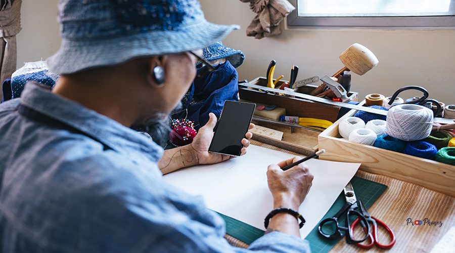 A man in blue clothes, hat and apron draw the pattern of his clothes on the paper with pencil and look at the reference on the internet by smartphone.
