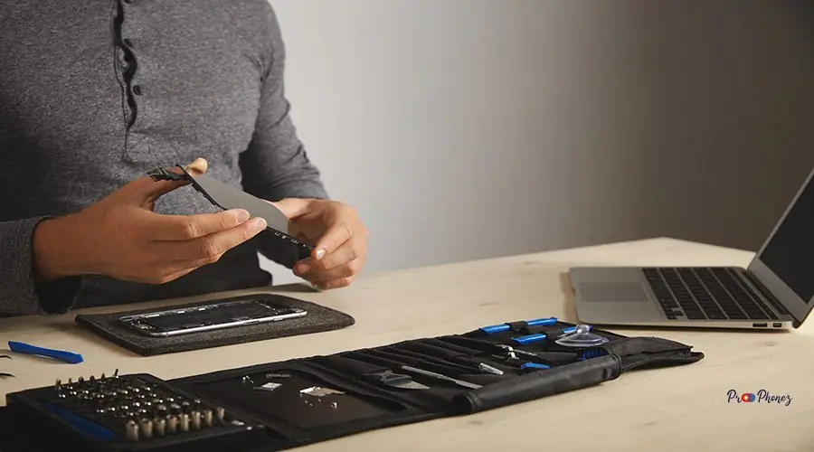 Computer and phone repairment service Master holds new screen for replacement above disassembled smartphone in his laboratory, tool kit with instruments and laptop in front of him on white table, space for your text on right
